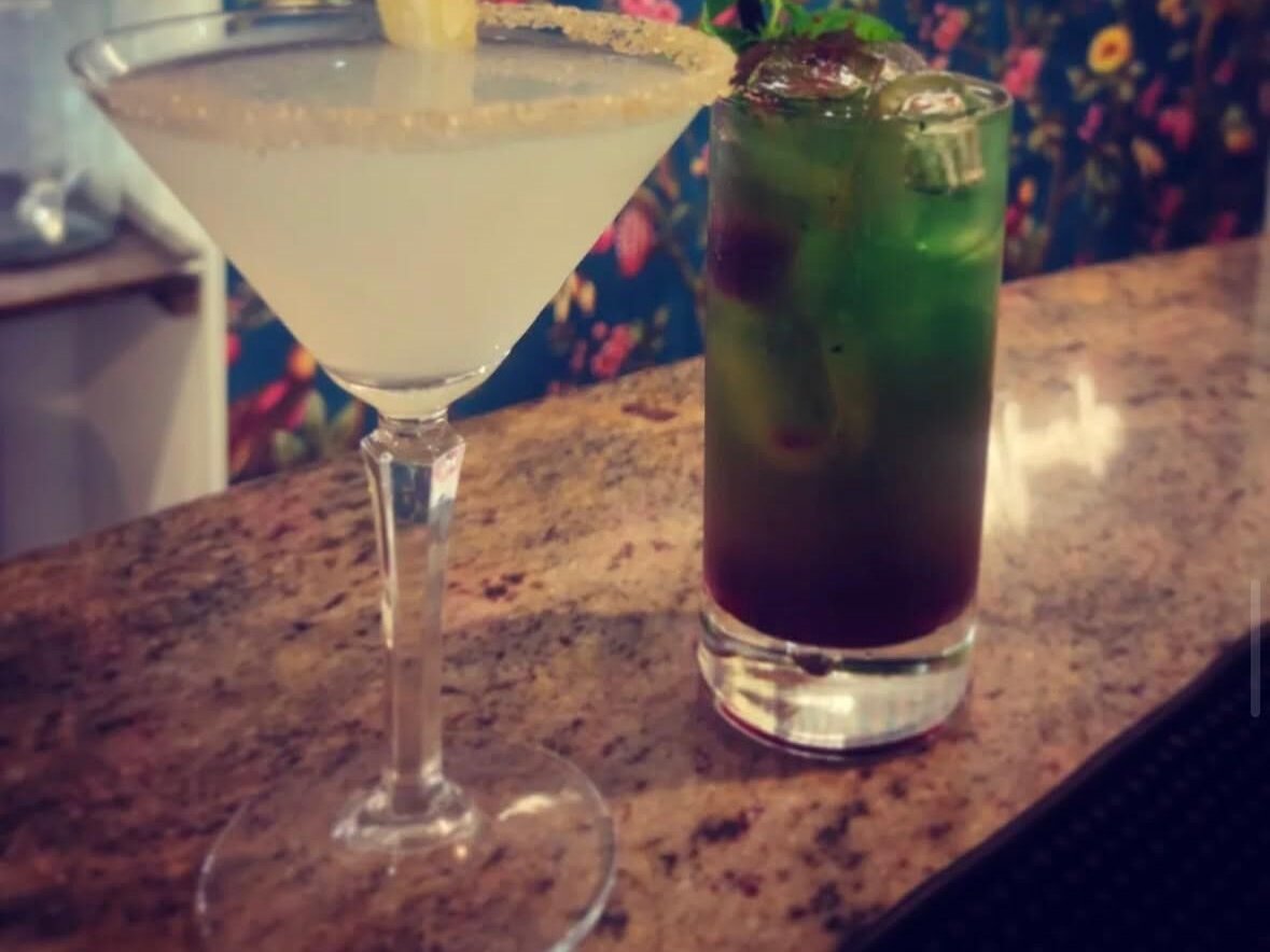 Coppa da martini con drink chiaro e bordo zuccherato accanto a un tumbler verde e viola con menta su bancone in granito